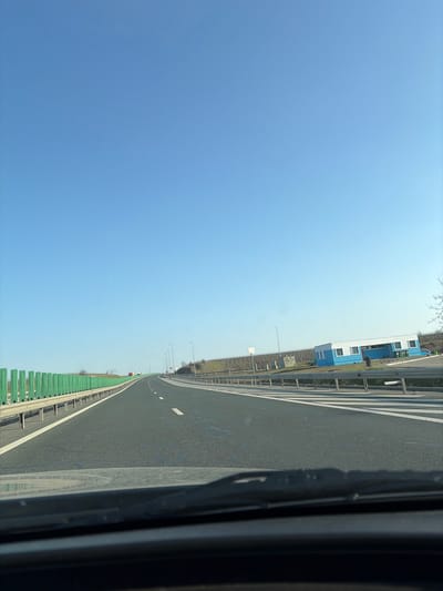 The image presents a static, forensic view captured from the interior of a vehicle, looking forward through a windshield onto a section of the A2 motorway near Cernavoda, Romania. The upper two-thirds of the frame are dominated by a clear, unclouded bright blue sky. In the immediate foreground, a dark vehicle dashboard and the lower edge of a windshield are visible, with a single black wiper blade partially obscuring the view on the lower right.
The central and midground feature a multi-lane asphalt highway, characterized by distinct white dashed lane markings and a solid white line delineating the right shoulder. The road appears to curve gently to the right as it recedes into the distance. On the left side of the highway, a series of vertically aligned green corrugated sound barriers are present, running parallel to the road and protected by a metallic guardrail.
On the right side of the highway, a metallic guardrail separates the main carriageway from an adjacent paved area, which could be a deceleration lane or service access, marked with diagonal white stripes. Beyond this paved area, a modular building stands on a slightly elevated paved surface. This building is predominantly blue with white trim around its windows and doors, and has a white roof. Several dark, cylindrical bins or containers are positioned near the building. The terrain behind the building ascends gently, covered by structured rows of dormant vegetation. Multiple street light poles line both sides of the motorway, extending into the distant perspective. No individuals or actively moving vehicles are present within the observable scene.