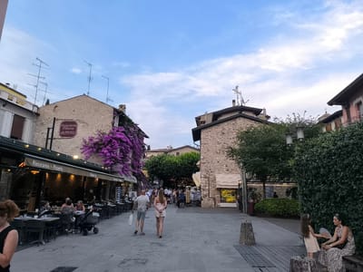 This image captures a street scene in Sirmione, Italy, during the late afternoon or early evening. The street is paved with cobblestones and lined with buildings on both sides. On the left, a restaurant with outdoor seating is visible, with patrons dining under a green awning. A vibrant cascade of purple bougainvillea adorns the building above the restaurant, partially obscuring a sign for "Ristorante Pizzeria VALENTINO."

Several people are present in the scene. A couple is walking away from the camera in the center of the street, while other individuals are seated at tables outside the restaurant. In the foreground on the right, a woman and a young girl are seated on a bench, with the girl holding a fan. The sky above is a pale blue with scattered white clouds. The overall atmosphere appears relaxed and leisurely, typical of a European town square or pedestrianized street.