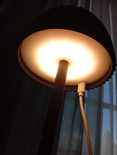 The image displays a close-up, low-angle shot of a modern desk lamp with its light on. The lamp features a wide, circular lampshade with a textured dark exterior and a smooth, off-white interior that diffuses the warm light emanating from the bulb. A slender, textured stand supports the lampshade, and a light-colored cable connects to the underside of the lampshade, presumably for power. The background is dark, with vertical lines suggestive of curtains or blinds, and a hint of blue light filtering through from the right side, indicating a window. The scene is devoid of people or any discernable activity. No specific location within Beyoğlu, Turkey is identifiable from the image's content.