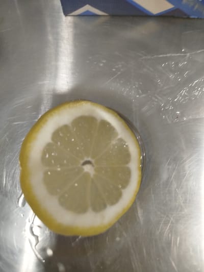 A close-up, overhead shot shows a slice of lemon on a metallic surface. The lemon slice is centered in the frame, with its yellow rind and pale yellow interior clearly visible. The surface appears to be a stainless steel countertop, which is brushed and shows several scratches. There are droplets of water on the surface, particularly around the lemon. In the upper portion of the image, a blue and white striped object is partially visible, suggesting it might be a box or packaging. The lighting is bright, creating reflections on the metallic surface. The location in Gelendzhik, Russia, is not discernible from the image content.