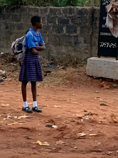 A young person in a blue shirt and plaid skirt stands near a brick wall. A sign reading "Swimming" is nearby. The person has a backpack and wears white socks with black shoes. The ground is reddish-brown dirt, littered with debris. The overall scene is outdoors.