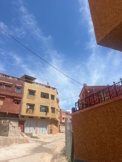 The image depicts a street view in Ouaouizert, Morocco. The perspective is looking upwards through a narrow alleyway between buildings. The sky is blue with scattered clouds. Buildings on either side are constructed of brown and yellow stucco with ornate wrought iron balconies. A power line stretches across the sky. At ground level, the street is paved and a stack of grey blocks is visible.