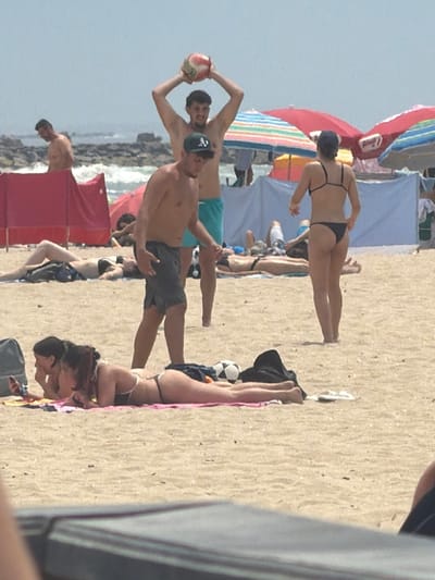 This image captures a lively beach scene in Espinho, Portugal, on a sunny day. In the foreground, two women are lounging on the sand, one with her arm extended and the other looking at a phone. Behind them, a group of people are enjoying the beach.
A man in the center is holding a volleyball above his head, as if preparing to serve. To his left, another man is standing and looking towards the sea. A woman in a black bikini is facing away from the camera, with her back to the viewer. In the background, colorful beach umbrellas and tents are visible, along with a glimpse of the ocean and rocky coastline. The sand is light brown and speckled with small shells and debris. The overall atmosphere is one of a typical summer day at the beach, with people relaxing and engaging in recreational activities.