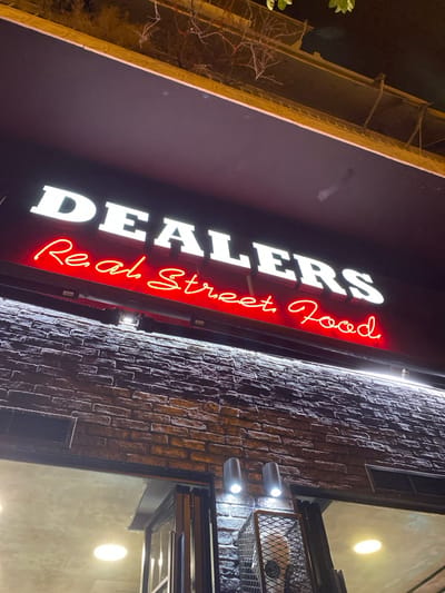 This media file presents a nighttime exterior view of a commercial establishment, specifically a food business, located in Koridallos, Greece. The scene is dominated by a brightly illuminated signboard, oriented diagonally across the image.
The prominent sign features the word "DEALERS" in large, white, capital letters with a slightly reflective or dimensional quality. Below this, in a distinct, red, neon-style cursive font, the text "Real Street Food" is displayed.
To the right of the sign, a textured brick wall runs vertically, showing various shades of brown and red due to the lighting. This wall is illuminated by several small, silver-colored spotlights that project focused beams of light, creating highlights and shadows on the brickwork. Further right, an opening or window reveals a lighter interior wall with at least two round, diffused light fixtures. A metal grate or basket-like structure is also partially visible in this area.
In the darker upper-left background, parts of another building or structure are visible against the night sky, appearing to have some metallic or light-colored framework, possibly scaffolding or architectural detail. Bare, leafless tree branches extend into the lower-left corner of the frame, suggesting a deciduous tree and potentially cooler weather.
The time of day is clearly night, evidenced by the dark sky and the strong reliance on artificial lighting to illuminate the sign and parts of the building. No people are visible in the image. The lighting creates a dynamic and inviting atmosphere for the business.
