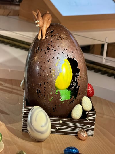 The image shows a large, elaborately decorated chocolate Easter egg display. The egg is mostly brown with a textured, hole-filled surface. Inside, there are yellow and green spheres, likely made of chocolate or candy. A small chocolate rabbit figurine is attached to the side of the egg. The egg is mounted on a dark chocolate stand with white chocolate drizzle decoration, and several smaller eggs are attached to the stand, including white, marble, and foil-wrapped eggs. A red, berry-shaped candy is also visible on top. The background is a light-colored wooden wall with vertical panels. The display is located at Thierry Mulhaupt, Illkirch-Graffenstaden, France.