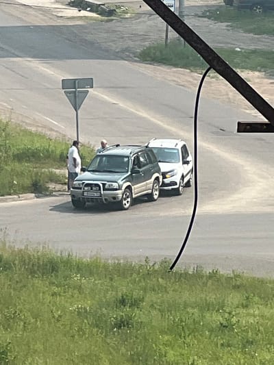 In Zheleznogorsk, Russia, two vehicles are parked on an asphalt road near a grassy verge. A dark green SUV is positioned in the foreground, facing slightly to the right. A white SUV is parked behind it, also facing to the right. Two individuals are visible standing near the green SUV. One, wearing a white shirt and dark pants, stands to the left of the vehicle, facing it. Another individual, wearing a light-colored shirt, is standing behind the green SUV, near the driver's side. A traffic sign with a triangular and rectangular component is situated on the left side of the road. The environment appears to be an urban or suburban setting with a road, grass, and a distant hazy background.