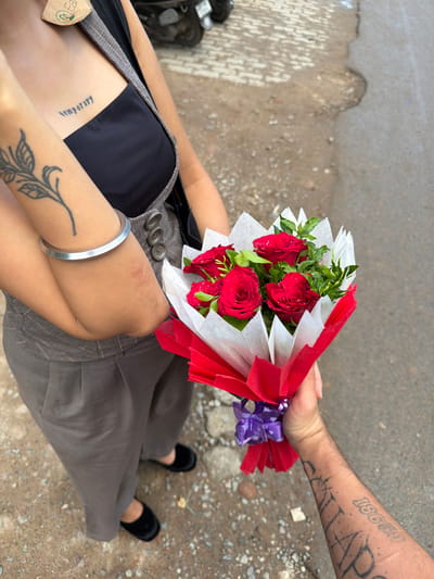A medium shot captures an individual offering a bouquet of red roses to another person in the city of Jajmau, India. The recipient, wearing a black camisole and gray overalls, has a tattoo of the word "temporary" on their chest and a floral tattoo on their left arm. They are also wearing a silver bangle on their left wrist and black velvet shoes. The giver's arm, adorned with several tattoos, is holding the bouquet, which is wrapped in white and red paper and tied with a purple ribbon. The background consists of a dirt and asphalt ground, with a hint of a parked motorcycle visible.