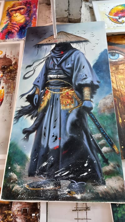 The image displays an oil painting of a samurai figure wearing a conical hat, a dark robe, and holding a katana with inscriptions on its blade. The figure is positioned against a backdrop of muted blues and greens, suggesting a natural setting. The painting is placed among other artworks in what appears to be an art gallery or studio, evidenced by the presence of other paintings on shelves and a display rack. This image was taken at Ban Ao Nang, Thailand.