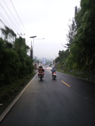 The image depicts a two-lane paved road at dusk or under heavily overcast conditions in Dawathang_Dorjibi_Kashingtsawa, Bhutan. The road surface appears wet, indicating recent precipitation. Two motorcycles are visible in the foreground, moving away from the viewer. The motorcycle on the left has one rider dressed in a brown jacket, with a possible passenger obscured behind them. The motorcycle on the right has a single rider wearing a light blue or grey jacket, and its red taillight is illuminated. A yellow broken centerline divides the road, and a solid white line marks the left shoulder. Lush green vegetation, including trees and dense bushes, lines both sides of the road, obscuring the terrain. Utility poles with power lines and at least one street lamp gantry are visible on the left side. In the blurred distance, multiple vehicle headlights and taillights are discernable, along with a large, indistinct billboard or sign above the road. The background shows hazy hills or mountains partially obscured by atmospheric conditions.