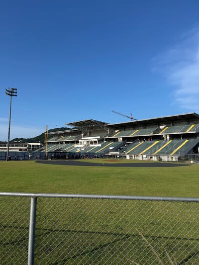 The image depicts the interior of a baseball stadium located in Curundú, Panama. The foreground is occupied by a grey chain-link fence with a horizontal metal top rail, positioned in front of a green grass outfield. The midground features the expansive, well-maintained green grass field, bordered by a dark warning track that curves along the perimeter. A group of approximately 20-30 individuals, indistinct in specific features, are visible near the dugout area along the first base line, appearing to be standing or casually gathered. Several more individuals are scattered throughout the multi-tiered stadium seating, which consists of green and yellow seats. The stadium structure includes overhead canopies providing shade to upper sections. A tall floodlight tower, equipped with multiple luminaires, stands prominently on the far left. In the upper right background, a large construction crane is visible above the stadium structure. The background also reveals a lush, vegetated hillside and several multi-story buildings on the far left. The sky is clear blue with sparse, wispy white clouds. The scene is illuminated by bright natural sunlight, casting distinct shadows and suggesting daytime conditions.