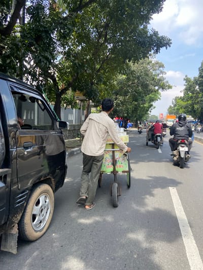 The image shows a man pushing a small food cart down a street in Kecamatan Cimahi Selatan, Indonesia (latitude: -6.909, longitude: 107.568). The man is wearing a light-colored button-up shirt, dark pants, and dark shoes. He is pushing the cart with his right hand. The cart is painted yellow and green with a white sign that reads "Kios Totos." There are two motorcycles driving in front of the man, one is black and one is blue. The black motorcycle has a rider wearing a dark helmet and black jacket. There is a black pickup truck parked on the right side of the frame. The street is paved and has a white line down the center. There are trees lining both sides of the street. The sky is bright blue with a few white clouds. The man is walking toward the camera. The scene is a busy street in a city, with vehicles and people moving in and out of frame.