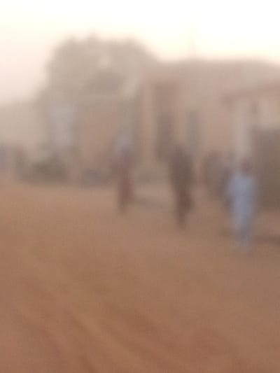 The image captures an outdoor scene in Rangaza, Nigeria, characterized by extreme blurriness and a pervasive hazy atmospheric condition. The foreground presents an unpaved, reddish-brown ground surface. In the middle distance, at least three human figures are present as indistinct vertical blurs. One figure on the right is distinguishable by lighter-colored attire, possibly blue or light grey. Other figures appear in muted, darker tones. The background contains faint outlines of structures, potentially buildings, in beige or light brown hues, along with vague shapes consistent with sparse vegetation, all heavily obscured by the prevalent haze. No specific objects, actions, or interactions are discernible due to the image's profound lack of clarity.