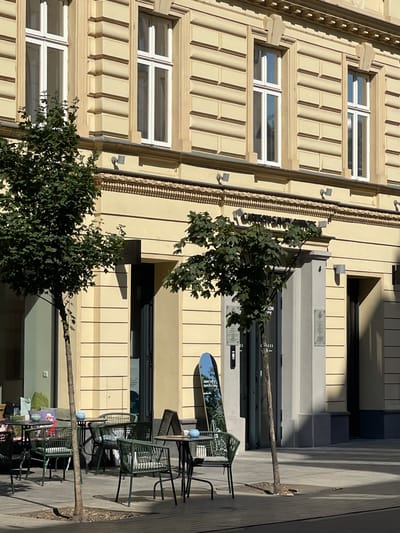 The image captures an outdoor seating area of a cafe located in Bratislava, Slovakia. The cafe is situated on the ground floor of a light yellow, multi-story building with ornate architectural detailing, including layered facade elements and decorative moldings above the windows. Several trees with green foliage are strategically placed around the seating area, providing shade.

The seating area consists of dark green metal tables and chairs. One table is set with blue bowls, and another has a dark-colored table runner. Cushions, some green and one with a patterned design, are visible on a bench or sofa in the background, suggesting the cafe is furnished for comfort. A slender tree trunk is prominently positioned in the foreground, with its shadow falling across the paved ground.

Various signs are visible. One is an A-frame sign placed on the pavement, and a tall, oval-shaped mirror leaning against a column displays text including "Summer is here. Season is a happy...". Above the entrance to the building, a dark sign reads "CARLTON SAVOY OFFICE". Two framed notices are mounted on a pillar near the entrance. The overall scene conveys a relaxed atmosphere on a sunny day, with ample natural light casting distinct shadows.