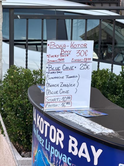 The image captures a close-up view of a sign advertising boat tours in Tivat, Montenegro, placed on a dark counter next to lush green foliage. The sign, written in red and black marker on white cardstock, lists two tours: "BOKA-KOTOR BAY" for 30€, running every day from 9:00 to 18:00, and "BLUE CAVE" for 25€, also running every day. The "BLUE CAVE" tour includes stops at (Submarine Tunnels), (Beach Žanjić), and (Blue Cave), with a start time of 10:00 and an end time of 16:00. Below the sign, a blue banner with the words "KOTOR BAY" and "Knez Lipovac" is partially visible, suggesting the location is a point of departure or information for boat excursions. The background shows a white vehicle and a black metal fence, hinting at an outdoor setting, likely near a harbor or tourist area in Tivat. A small piece of paper with handwritten text and a phone number, possibly for booking, is taped to the counter.