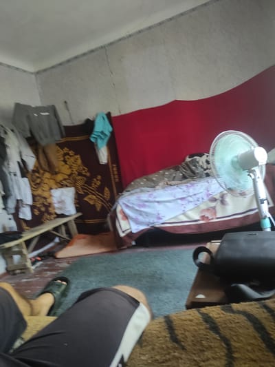 The image depicts an indoor scene, likely a bedroom or living area, with a cluttered and lived-in appearance. In the foreground, a person's legs are visible, wearing dark shorts and one black sandal. The room contains various items of clothing hanging on what appears to be a rack or wall, including a gray sweatshirt and other garments. A patterned rug or blanket with a floral design is draped over a surface, possibly a bed. There is a wooden bench or stool against a wall. A standing fan with a light blue base and grill is positioned on the right side of the frame, near a red fabric backdrop. A dark handbag or briefcase is visible on a small wooden surface. The walls are textured and appear to have some cracks. The floor is covered by a dark green carpet. The overall impression is of a modest, perhaps somewhat unkempt, domestic space. No specific landmarks or indications of the city of Ungheni, Moldova, are present within the image itself.