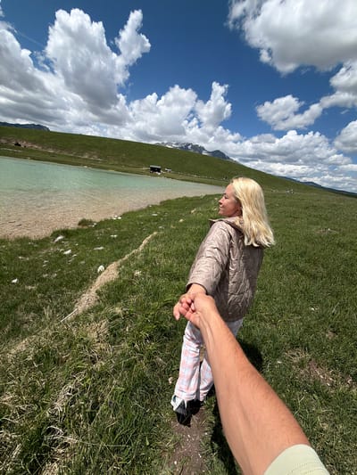 The image depicts a first-person perspective in Javorje, Montenegro, featuring a person extending their right arm to hold hands with a blonde woman, leading her through a grassy landscape. The arm in the foreground, belonging to the photographer, is light-skinned with visible hair and a light green sleeve. The woman, appearing in her 30s-40s, has long, wavy blonde hair and wears a light brown quilted jacket and light-colored plaid pants. She looks towards the right side of the frame, away from the camera, with a neutral expression.

The setting is a high-altitude natural environment under a partly cloudy blue sky. A tranquil, light turquoise lake or pond occupies the middle ground on the left, with visible light-colored sediment and stones along its shallow edge. The surrounding terrain consists of verdant grassy hills, some with subtle dirt paths. In the far distance, snow-capped mountains are visible. On the opposite side of the lake, a small dark-colored cabin, a white vehicle, and a yellow object (possibly a boat) are discernible.