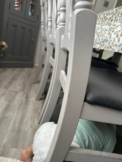 An extreme low-angle, medium shot captures the legs of a baby wearing a light-colored onesie patterned with elephants, tucked under a light grey chair. The baby is positioned on a pale wooden floor. The chair, made of painted wood, has a black seat and is part of a set of dining chairs. A table runner with a gold and grey holly pattern is visible on the table behind the chairs. In the background, a dark grey cabinet with mirrored doors is present. The scene appears to be a domestic interior, likely a dining room, in Macclesfield, United Kingdom.
