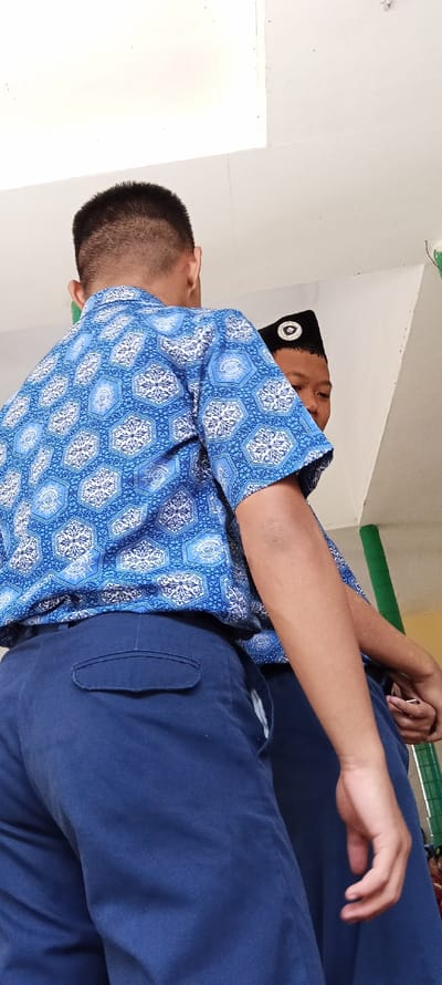 The image shows two young men standing in a hallway, likely at a school. The man on the left, in the foreground, is wearing a blue shirt with white geometric patterns and blue pants. The man on the right is wearing a black cap, a white shirt, and blue pants. The hallway walls are painted white and there is a green painted column in the background. Both men are looking towards the camera. The scene is bright and well-lit. The location is at the coordinates -7.065, 107.551, which is in Cangkuang, Indonesia.