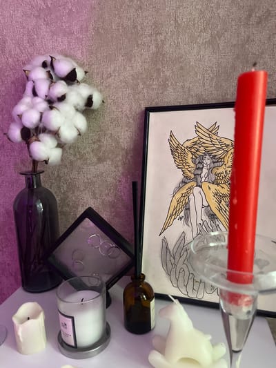 The media file displays a still life arrangement with a focus on various decorative and symbolic objects. In the foreground, a cluster of white cotton bolls rests on a textured, possibly fabric, surface that is illuminated with a soft purple light. To the right of the cotton, a dark-colored glass bottle lies horizontally, its opening plugged with a stem. Above and behind this bottle, a small, black framed display case contains several silver rings.
Further back, a framed drawing is visible, featuring stylized figures with wings, rendered in black ink with yellow accents. Partially obscuring the drawing is a bright red candle held within a clear glass holder. To the right of the drawing, several white candles are arranged, one in a glass jar and another as a smaller, partially melted pillar. A small amber glass bottle, possibly an oil or perfume dispenser, is also present. The overall setting appears to be indoors, possibly a tabletop or shelf display, with the textured background and artificial lighting contributing to an ambient, perhaps decorative, atmosphere. No specific time of day or weather conditions are discernible. The visible text on the candle in the glass jar is too small and blurry to be read. The context provided suggests the location is Samara, Russia.