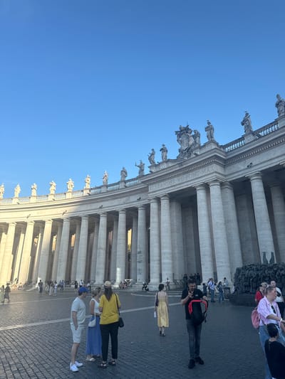 The image shows a partial view of St. Peter's Square in Vatican City, Rome. A section of the colonnade, constructed by Bernini, is prominently visible with its Doric columns and statues lining the top. People are walking around the square. One man is photographing a woman in a yellow dress. In the background are other tourists. The sky is clear and blue. The colonnade provides shade and shadows, indicating a sunny day.