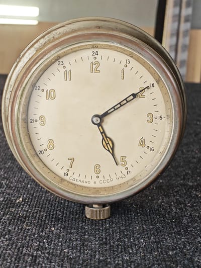 A close-up, eye-level shot shows a vintage Soviet-era clock with a cream-colored face, gold Arabic numerals, and black hands. The clock has a worn, metallic bezel and is set on a dark, textured surface. The time displayed on the clock is approximately 1:59:30. The phrase "СДЕЛАНО В СССР 443" is visible on the lower part of the clock face, indicating it was made in the USSR. The background is out of focus, showing a blurry interior with hints of lighting fixtures and shelving. The specific location within Ждановіцкі сельскі Савет, Belarus, is not discernible from the visual information provided in the image.