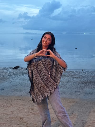 A young woman stands on a beach in Baan Sri Thanu, Thailand, making a heart shape with her hands. She has long, dark hair and is wearing light jeans and a poncho-style top with fringe. The beach is sandy and meets the water of a bay under a cloudy, blue sky. The scene is likely taken at dusk.
