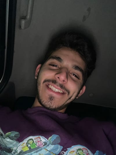 A young man smiles at the camera from the passenger seat of a car. He has dark curly hair, brown eyes, and a trimmed beard. The car interior is dimly lit. He wears a purple sweatshirt with a cartoon design. The roof of the car and a grab handle are visible above his head.