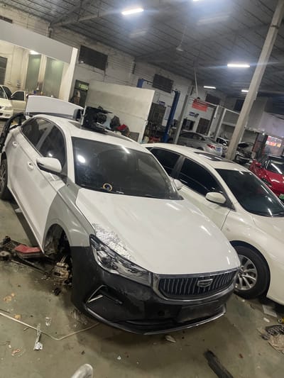 The image depicts the interior of a car repair shop in Panyu District, China. A white sedan, partially disassembled with its front bumper removed and front wheel exposed, is the primary subject. The car is situated on a lift or jack. To the right of the white sedan, another white sedan is parked, and a red sedan is visible further in the background. The environment is a dimly lit industrial space with exposed ceiling beams, fluorescent lighting, and various tools and car parts scattered on the floor. The overall scene suggests an active auto repair facility.