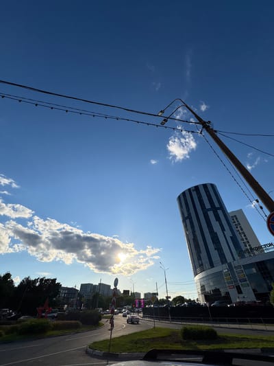 The image depicts a bright, sunny day in Mytishchi, Russia, with a clear blue sky and scattered white clouds. The sun is shining through the clouds, creating a lens flare effect. In the foreground, a thick cable runs diagonally across the frame, supported by utility poles and strung with small lights. In the middle ground, a modern, curved building with a glass facade and horizontal dark bands dominates the scene. Adjacent to it, another taller building with a similar facade is visible. A road curves in the foreground, bordered by a grassy verge and some trees. Traffic is visible on the road, indicating an urban or suburban setting. The overall mood is one of a bright, clear day in an urban environment. The text "КОЕСТОК" is visible on the taller building. The perspective is from a slightly elevated position looking upwards and outwards.