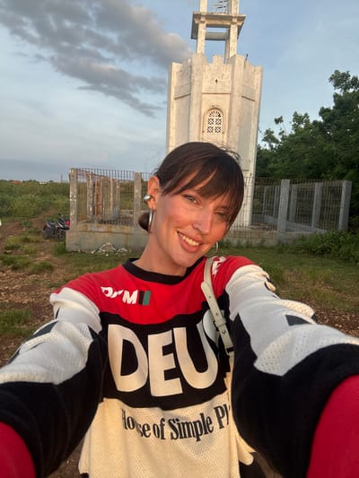 The image captures a female subject taking a selfie in Uluwatu, Indonesia, under soft, natural lighting conditions. The subject smiles directly at the camera, revealing dark brown hair with bangs, pulled back into a low bun. She has a nose ring, a hoop earring on her visible left ear, a silver ear cuff on her upper left ear, and another hoop earring partially visible on the right. She wears a long-sleeved jersey-style shirt with red, black, and white panels, featuring "DEUS House of Simple Pleasures" printed prominently on the front. A light-colored shoulder bag strap crosses her right shoulder.
In the background, a weathered white concrete tower stands centrally. This structure features a crenellated top, an arched window with a decorative grate, and an open top level with exposed concrete beams, appearing disused or unfinished. Surrounding the base of the tower is a dilapidated perimeter fence made of concrete posts and metal mesh. The ground in the foreground and to the left is unpaved and dry, with scattered green scrub vegetation. A dark-colored scooter is parked on a dirt track to the far left. The sky above is light blue with scattered white clouds.