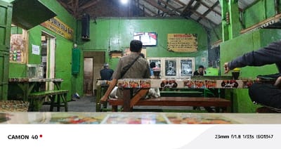 The scene takes place inside a brightly lit, open-air restaurant in Pasuruan, Indonesia, located at coordinates -7.663 latitude and 112.906 longitude. The walls are painted a vibrant green. The restaurant's interior features wooden tables and benches, and a flat-screen television mounted on the wall displaying a news broadcast.
A man with short black hair, wearing a brown T-shirt and black backpack, is seated at a table in the center of the scene. He is looking at the television, and has a cup of coffee in front of him. There is a poster on the wall behind him with the word "MENGHARGAI" written on it.
To the left of him, a second man, wearing a gray shirt, is seated at a table and appears to be looking towards the man with a cup of coffee in front of him. There are several tables with seating in the scene. At least two other people can be seen sitting at a table on the right side of the scene.
The restaurant has a wooden roof that can be seen in the background. The floor is a mix of concrete and tile. The camera is situated at a low angle, pointing towards the center of the scene. The lighting in the restaurant is bright and warm. The image appears to have been taken with a phone camera.