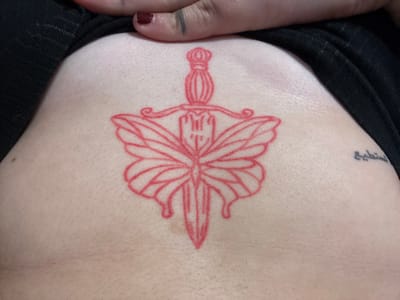 A close-up photograph shows a tattoo on a person's upper chest/sternum area. The tattoo is a symmetrical design, primarily red in color, featuring a dagger or sword as its central element. Surrounding the dagger are wing-like shapes, resembling a butterfly or moth. A small portion of a black garment is visible at the bottom and left edge of the frame. A finger with red nail polish is visible on the bottom edge of the picture. On the upper left, just above the neckline, is a small tattoo of Arabic script. The background is the person's skin. The location of the picture is not detectable from the image. Therefore, Kuklen, Bulgaria, is not verifiable.