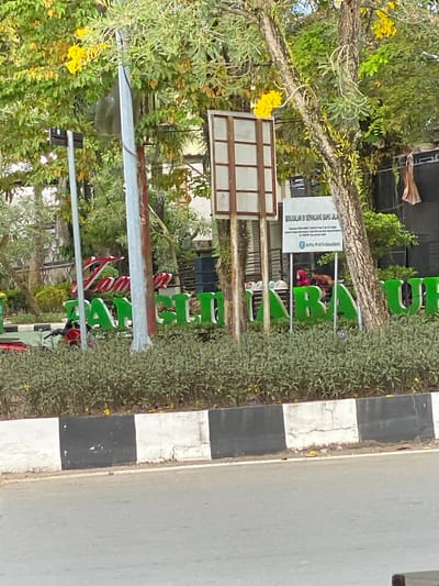 The image depicts a street scene in Banjarbaru, Indonesia, with a focus on a large green sign spelling out "Banjarmasin" in prominent lettering. The sign is positioned behind a low wall painted with alternating black and white blocks. Lush green foliage, including trees with yellow flowers, forms a backdrop for the sign. To the left of the sign, a red and white striped sign is partially visible. In front of the green sign, two individuals are seen; one wearing a red hijab and another in a brown shirt. A motorcycle with a red seat is parked near the green sign. A light pole with some signage is visible to the left of the scene. In the upper right quadrant of the image, a white rectangular sign with Indonesian text is mounted on a pole, and behind it, dark structures resembling speakers are present. The overall environment appears to be an outdoor public space, likely a park or a designated area along a road.