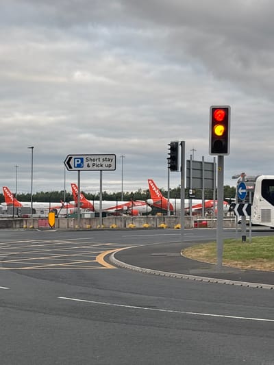 The image depicts an outdoor scene at what appears to be an airport, likely in Manchester, United Kingdom, under an overcast sky. In the foreground, an asphalt road with distinct white and yellow markings, including a yellow box junction pattern, is visible.
On the left side of the frame, a traffic light pole stands tall, displaying a red light at the top and an amber light in the middle. Below this, a smaller, darker traffic light unit is present. Adjacent to the traffic lights, a prominent white sign with a blue border is visible, featuring a "P3" symbol and text that reads "Short stay & Pick up", accompanied by an arrow pointing to the left.
Beyond these elements, several orange and white EasyJet airplanes are parked in a row, with their tailfins and parts of their fuselages clearly visible. A green treeline separates the planes from the distant cloudy sky. On the far right, a white airport bus or service vehicle is partially visible, displaying a blue downward-pointing arrow on its front. The scene is static, depicting an airport's ground operations area during daylight hours, characterized by infrastructure and parked aircraft.