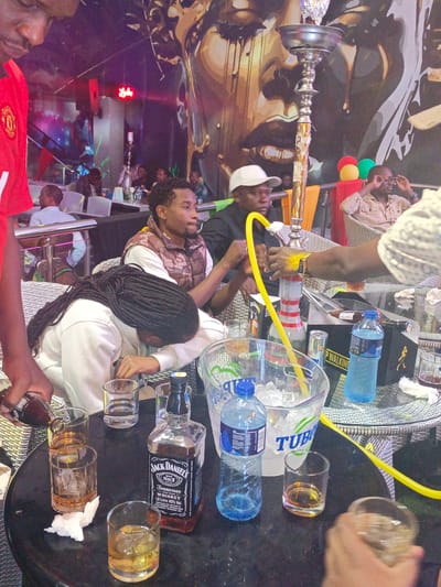 The scene depicts a group of people at a table in an indoor establishment, likely a bar or lounge, in the Syokimau-Mulolongo ward, Kenya. A hookah stands on the table, attended to by a person wearing a yellow-gold wristwatch. A bottle of Jack Daniel's whiskey sits prominently on the table, accompanied by several partially filled glasses, suggesting drinking activity. A bottle of Coca-cola is also visible, being poured into one of the glasses. A woman with braided hair is seated at the table, with her head lowered. Two men are seated across from her and the hookah attendant, one wearing a vest. Another person is seen wearing a red Manchester United jersey. An ice bucket, with a "TUBO" label, holds ice, and water bottles are also present. The background features a mural of a face on the wall, colorful lighting, and other patrons seated at tables.