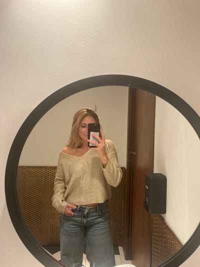 A female subject is captured in a mirror selfie, standing indoors. She has blonde hair and wears a light-gold, V-neck, long-sleeved sweater and light-wash blue jeans; her left hand is placed in a jean pocket. The subject holds a black smartphone with a white card in the back, partially obscuring her face, to photograph her reflection in a large circular mirror with a thick dark frame. The reflected environment includes a gold-colored wall panel with a diamond-grid texture, a dark brown wooden door, a white wall, and a black wall-mounted dispenser. A black and white checkerboard tiled floor is visible at the bottom of the reflection. The specific location within Santander, Spain, is not discernible from the visual evidence.