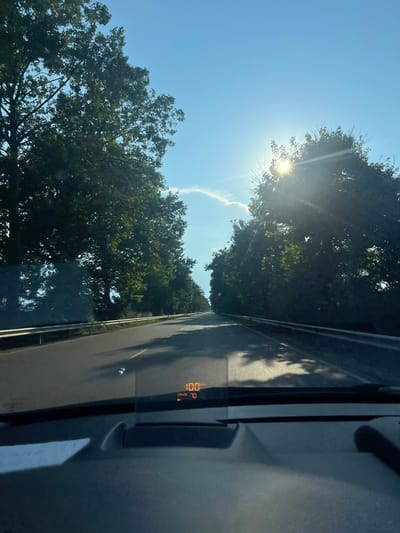 The image depicts a scene from inside a vehicle looking outwards. The sun is bright and high in a clear blue sky, casting rays through the green leaves of trees that line the road. The trees suggest a rural or suburban setting, with lush foliage indicating a warm season. The road surface is visible, with tire tracks and a faint white line marking the lane.

On the interior of the vehicle, a digital display shows "100" which likely represents the speed in km/h, and "CRUISE 70" indicating the cruise control is set to 70 km/h. The time of day appears to be midday or early afternoon due to the bright sunlight and high sun angle. The weather is sunny and clear, with a few wispy clouds in the distance.

The perspective suggests the photo was taken while the vehicle was in motion. The overall mood is one of a pleasant journey on a sunny day. The location context is Orizari, Bulgaria.