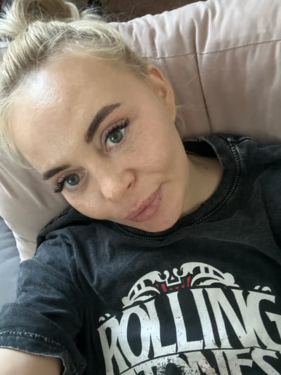 A light-skinned woman with blonde hair and grey-blue eyes is lying down. She appears to be resting on a pillow, and she is wearing a dark grey t-shirt with a white design. The location is Высокий, Russia.