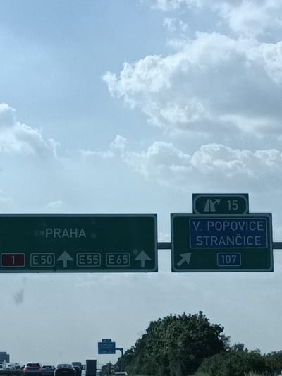 This image shows a highway scene on a cloudy day, likely in the Všechromy, Czechia area. The primary subjects are road signs directing traffic.
On the left, a green highway sign indicates "PRAHA" (Prague) with route numbers "1", "E50", "E55", and "E65", all pointing straight ahead.
To the right, a smaller sign above a green destination sign shows a symbol and the number "15". The green sign below it indicates destinations "V. POPOVICE" and "STRANČICE", with route number "107" and an arrow pointing right.
Below the signs, several cars are visible on the road, suggesting the image was taken from a moving vehicle. The sky is filled with large, white clouds against a light blue background, indicating daytime with overcast conditions. The overall scene depicts a typical European highway environment.