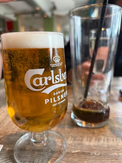 The scene depicts an indoor setting within an establishment in Felixstowe, United Kingdom. In the foreground, a stemmed Carlsberg glass is approximately three-quarters full of golden-amber beer with a head of white foam, resting on a light-colored wooden table exhibiting visible grain. The glass features the white Carlsberg logo, a crown emblem, and text including "BY APPOINTMENT TO THE ROYAL DANISH COURT," "DANISH PILSNER," and "1847 ONWARDS." To the right of the beer glass, a tall, clear, empty glass stands on the same table. This second glass contains a small amount of dark liquid residue, a lime wedge at the bottom, and a black straw extending into the residue. The background is out of focus, revealing blurred elements consistent with an indoor public space, such as indistinct walls and reflections, but no identifiable individuals or specific actions are discernible.