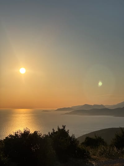 The image depicts a sunset over the sea, as viewed from a hilltop overlooking Kovačko Polje, Montenegro. The sun, a bright, glowing orb, is positioned in the upper left quadrant of the frame, casting a golden sheen across the water. The sea itself is calm, reflecting the warm hues of the sunset, with distant, silhouetted mountains forming a layered backdrop along the coastline. In the foreground, the dark silhouettes of trees and vegetation create a stark contrast against the luminous sky and sea, suggesting a viewpoint from a densely vegetated area. The sky transitions from a pale yellow near the horizon to a soft blue at the top of the frame. A second, fainter circular lens flare is visible in the upper right portion of the sky. There are no people or animals visible in the image. The overall scene is serene and natural, dominated by the elements of sky, sea, and land at dusk.