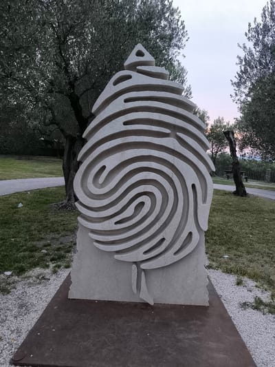 The image depicts a large, light-colored stone sculpture resembling a fingerprint, set in an outdoor park-like setting. The sculpture is the central focus, with a base that appears to be made of darker metal. The sculpture itself has a rough, textured surface and is carved with concentric lines that mimic fingerprint ridges, tapering to a triangular point at the top. The base of the sculpture is rectangular and dark.

Behind the sculpture, there are several olive trees with dense foliage, their trunks visible and gnarled. A paved pathway runs through the grassy area. In the background, beyond a low fence, a wooden bench is visible next to another tree. The sky above is a soft gradient of pale blue and pink, suggesting either dawn or dusk.

The overall impression is one of an artistic installation in a natural environment. The sculpture's form suggests themes of identity, nature, or perhaps connection. The lighting is muted, contributing to a serene atmosphere.

This sculpture is located in the "Parco di Villa Cortine" in Sirmione, Italy, specifically near the Grotte di Catullo.