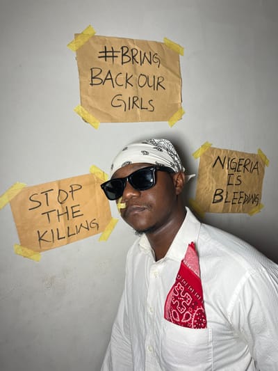 A person in a white shirt and bandana poses next to brown paper signs taped to a wall. The signs display messages: "#Bring Back Our Girls," "Nigeria Is Bleeding," and "Stop The Killing." The person wears sunglasses and a red bandana is tucked into their shirt pocket. The image is oriented on its side, giving the impression the person is lying down or the photo was rotated.