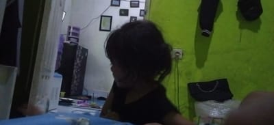 The image captures a dimly lit indoor scene, likely a room in Banjaran, Indonesia, based on the context provided. In the foreground, a young child with dark hair is the primary subject, facing to the right and slightly downwards, obscuring their facial features. The child appears to be wearing a dark-colored top.
To the left of the child, a tall white appliance, possibly a water dispenser or cooler, is visible. Further left, a dark cabinet with decorative patterns is present. In the background, on a white wall, several framed pictures are displayed in a cluster, along with a single framed picture on a separate wall to the left. A purple plastic item, possibly a water bottle, sits atop the dark cabinet.
On the right side of the image, a bright green wall is prominent. An electrical outlet with a cord is visible on this wall. Below the outlet, a black handbag with a chain strap rests on top of a white, plastic-wrapped object. The overall impression is of a casual, lived-in domestic environment.