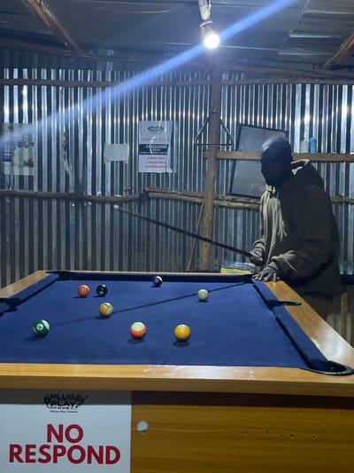 The image depicts an indoor billiards hall in Wumat, Nigeria. A person wearing a dark hooded sweatshirt and a dark baseball cap is playing pool, holding a cue stick and aiming at the balls on the table. The room has corrugated metal walls and a wooden ceiling with exposed beams. A bright light bulb hangs from the ceiling, illuminating the scene. Several pool balls are scattered across the blue felt-covered table. Posters and a triangle rack are visible on the walls. A sign at the bottom of the image reads "PLUG O PLAY HUB" and "NO RESPOND".