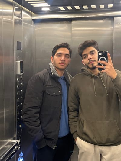Two young adult males are depicted inside a brushed stainless steel elevator, located within Kafr Sousa Municipality, Syria. The elevator interior features reflective walls, a control panel with numerous circular buttons and a vertical digital display on the left side, and recessed rectangular and linear light fixtures on the ceiling. The male on the viewer's left has short dark hair and a light beard, wearing a dark jacket with a light-colored, possibly faux-fur, collar lining over a blue long-sleeved shirt. His left hand is partially obscured, possibly in a pocket. The male on the viewer's right has curly dark hair and a light beard, wearing an olive-green hooded sweatshirt with a front pouch pocket and light-colored trousers. He is holding a dark-colored smartphone, equipped with a white circular accessory on its back, in his right hand, angled to capture their reflection in a mirror selfie. A dark ring is visible on his left ring finger. Both individuals are looking directly towards the camera's perspective.