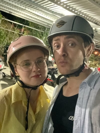 A selfie-style photograph, rotated counter-clockwise, shows two individuals wearing helmets. The person on top wears a gray helmet with a white stripe and a "Napoli" emblem. They have light skin, dark hair, and are making a pouting facial expression. The person below wears a pink helmet, glasses, and a yellow shirt. Both are secured with black helmet straps. The background shows glimpses of other motorcycles, vegetation, and a white metal fence. Based on the metadata, this photo was taken in Diên Khánh, Vietnam.