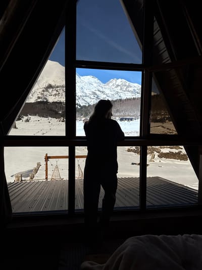 The image depicts an indoor perspective from a dwelling in Virak, Montenegro, looking out through a large, multi-pane window onto a snow-covered winter landscape. A solitary, silhouetted individual, possessing long hair and an average build, stands facing away from the camera, positioned centrally within the lower half of the main window frame. The person's hands are resting on the lower horizontal window beam, indicating observation of the external environment.

The interior of the room is dimly lit, revealing dark window frames and a sloped, possibly wooden, ceiling structure on the sides and top, characteristic of an A-frame or attic space. Dark curtains are pulled back on the left side. A glimpse of a light-colored blanket is visible in the bottom right corner, suggesting a bed or resting area.

Outside the window, a corrugated metal roof is visible in the immediate foreground. Beyond this roof, a vast expanse of snow-covered ground extends. Two hanging wicker chairs are suspended from a wooden crossbeam structure in the midground, situated on the snow. Further into the midground, several small, dark-roofed cabins are scattered across the snow-covered terrain, interspersed with bare deciduous trees and some coniferous vegetation. The background is dominated by a range of rugged, snow-capped mountains under a clear, bright blue sky, indicative of a sunny winter day.