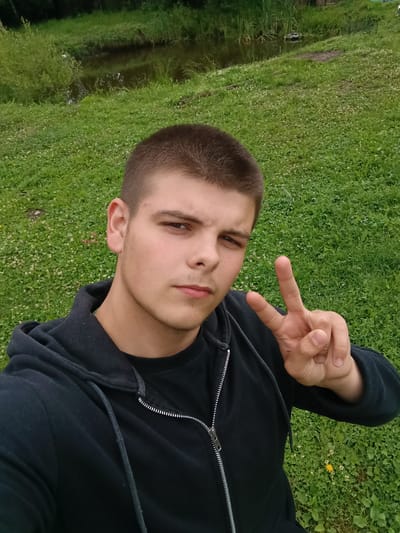 In this selfie, a young man with a buzz cut and a black hoodie is captured outdoors in a grassy area. He is looking directly at the camera and holding up a peace sign with his right hand. The background suggests a natural setting with a body of water, possibly a pond or a small river, surrounded by lush green grass and some reeds or tall plants. The overall environment appears to be rural or a park-like area. There are no specific indicators to place this image in Andreyevo, Russia.