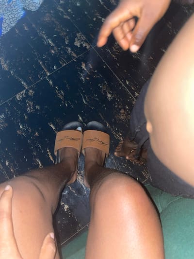 A low-angle shot captures two pairs of feet and part of a hand against a dark, glossy tile floor. One individual wears black trousers, revealing bare feet with visible toes. Another person dons tan-colored sandals with black soles, brandishing designer branding. The dark floor exhibits a marble-like texture, with variations in black and brown hues. Part of a hand appears near the floor, with part of the body out of focus.